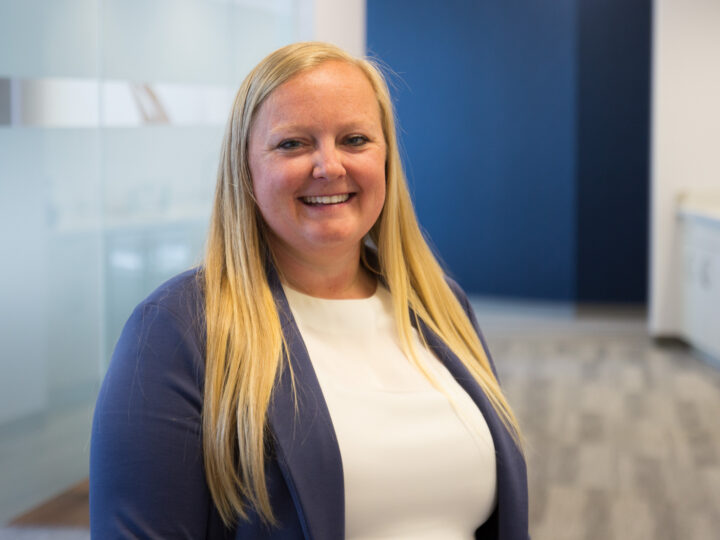 Smiling blonde woman in a navy blazer and white top, standing in a glass-walled office environment.