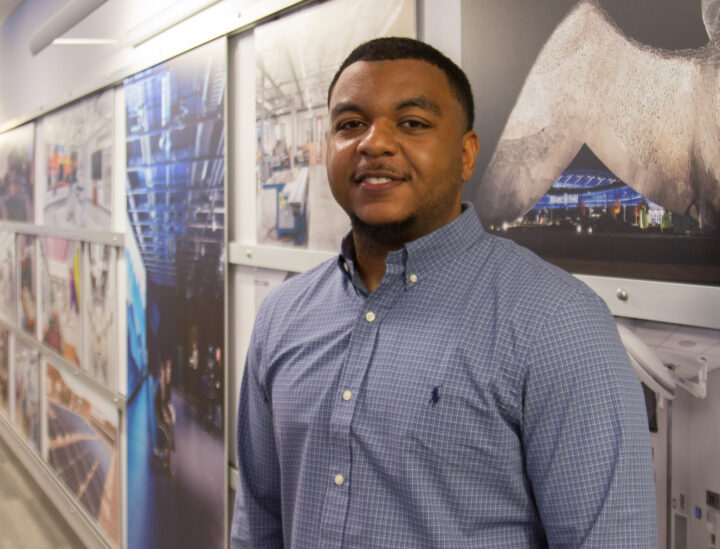 Portrait of a man in a blue checkered shirt standing beside wall-mounted posters inside a gallery or hallway, looking at the camera and smiling.