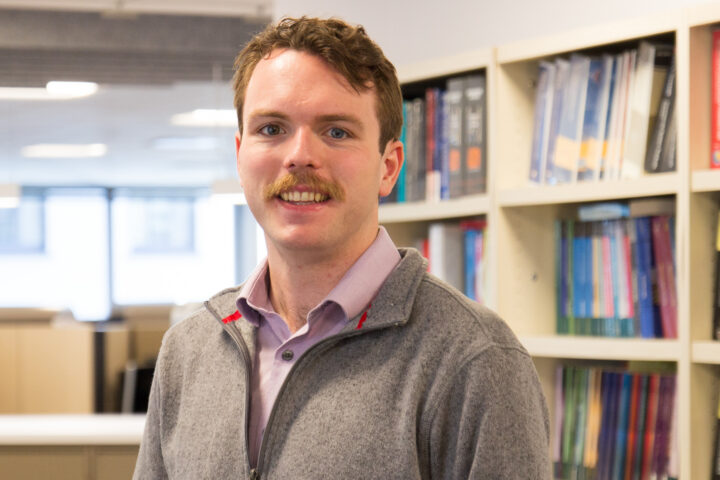 Smiling man with mustache in a library, bookshelves behind him.