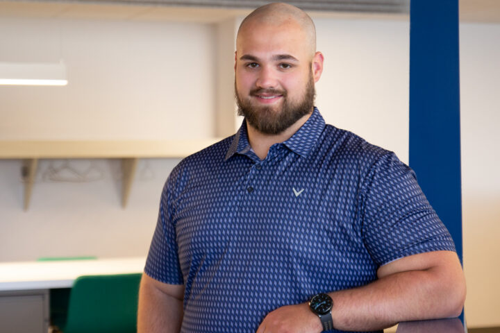 Portrait of a man with a shaved head and beard, wearing a blue patterned shirt in an indoor office space, arms crossed and smiling slightly.