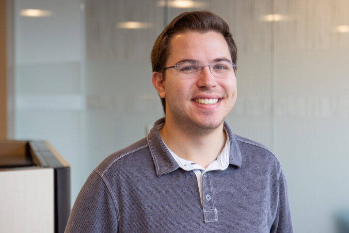 Smiling man with glasses in a blue-gray shirt standing in an office setting.