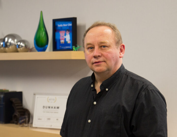 Man in a checkered shirt standing in an office, partial view of his head and shoulders with shelves and a framed poster behind him.