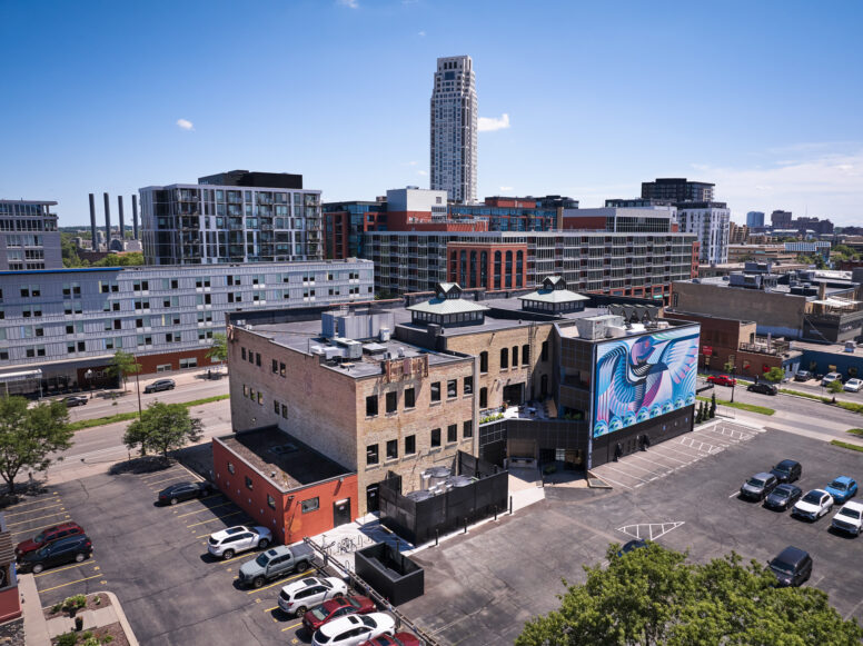 City skyline with a tall central skyscraper, surrounding modern buildings, and a blue mural on a lower wall beneath a clear blue sky.