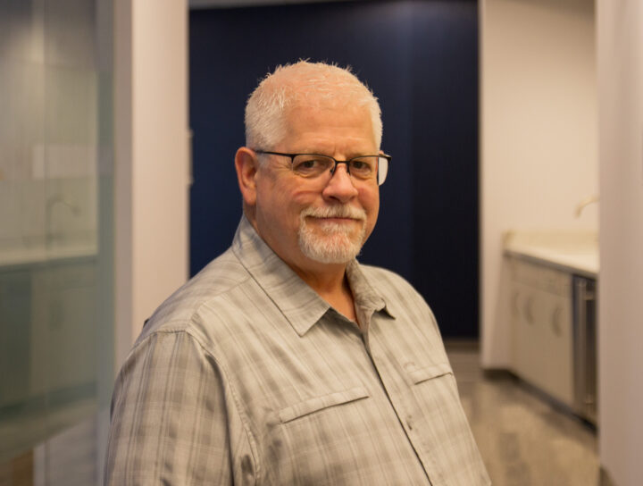 Older man with gray hair and a beard wearing glasses, in a plaid shirt, standing in an office hallway with glass and blue panels behind him.